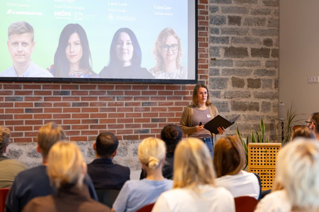 Piret Seeman stands before a seated audience in a brick-walled room, holding an open folder, with a slide of the event speakers projected behind her.