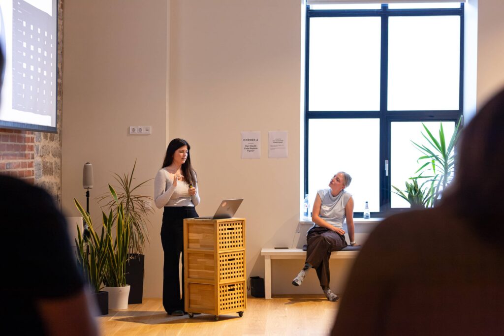 Laura Ader presents from a laptop on a wooden lectern beside a tall window, with an audience member sitting on the window bench.
