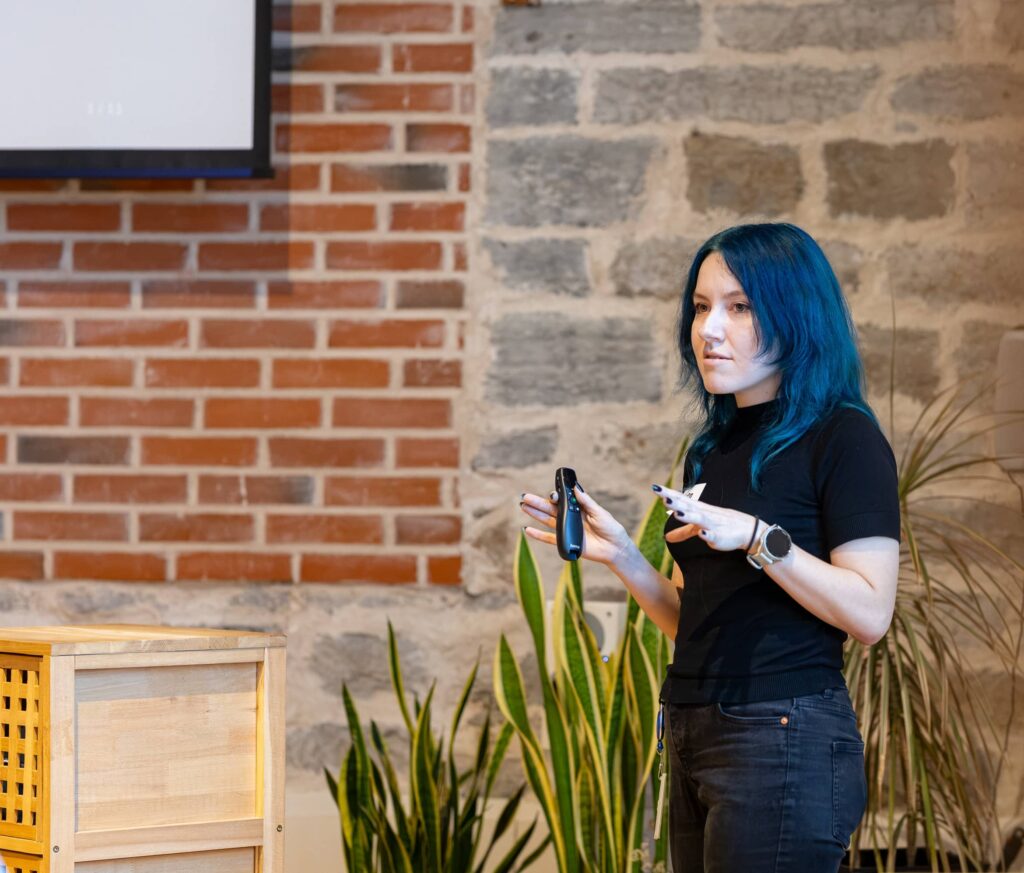 Laura Leon, with long blue hair, speaks in front of a brick and stone wall, holding a presentation clicker, with the projection screen at the edge of the frame.